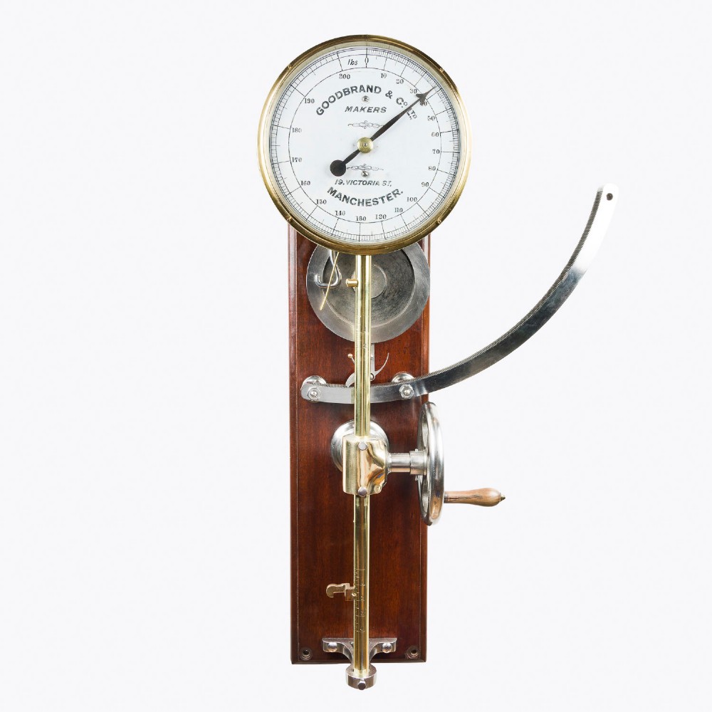A wall-mounted antique Goodbrand & Co brass and steel yarn testing instrument mounted on a rectangular mahogany board with a circular dial scale.