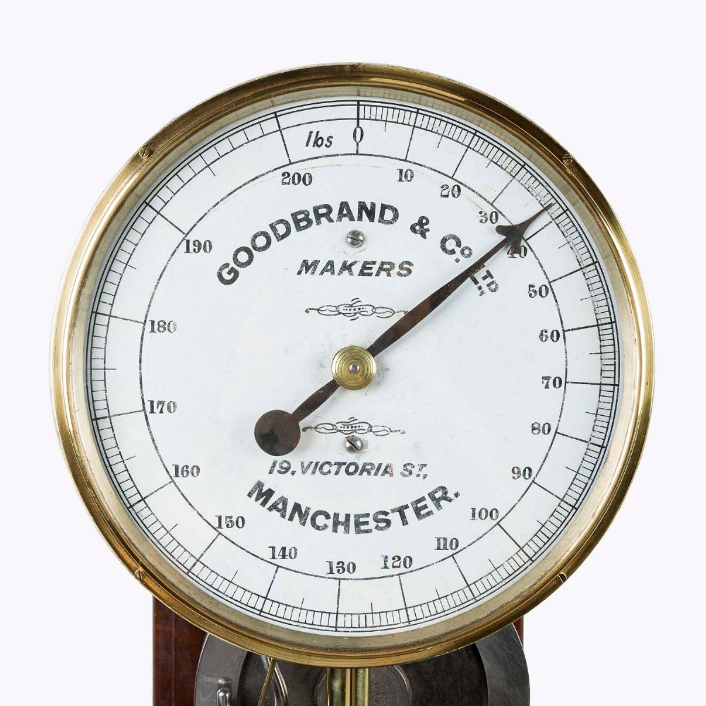 A wall-mounted antique Goodbrand & Co brass and steel yarn testing instrument mounted on a rectangular mahogany board with a circular dial scale. View II