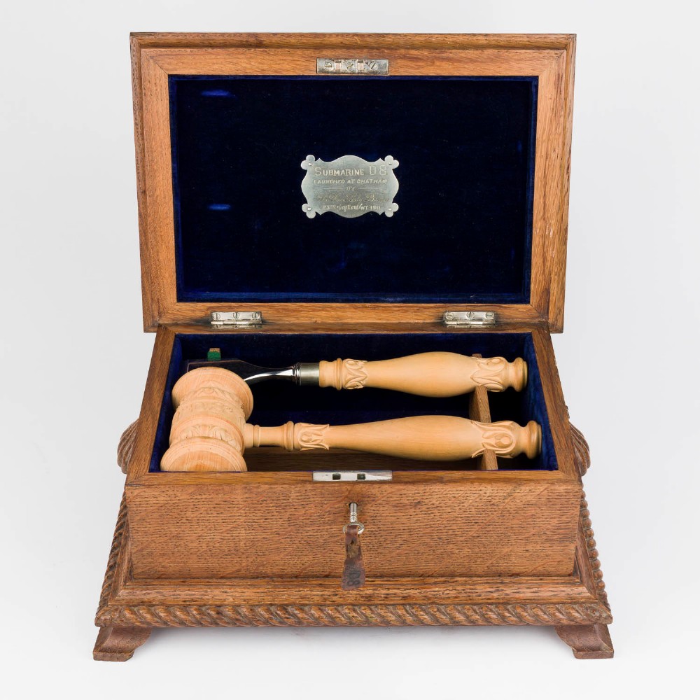 An open Edwardian carved oak presentation casket lined in navy blue velvet, containing a carved wooden ceremonial mallet and chisel set with an engraved silver plaque.