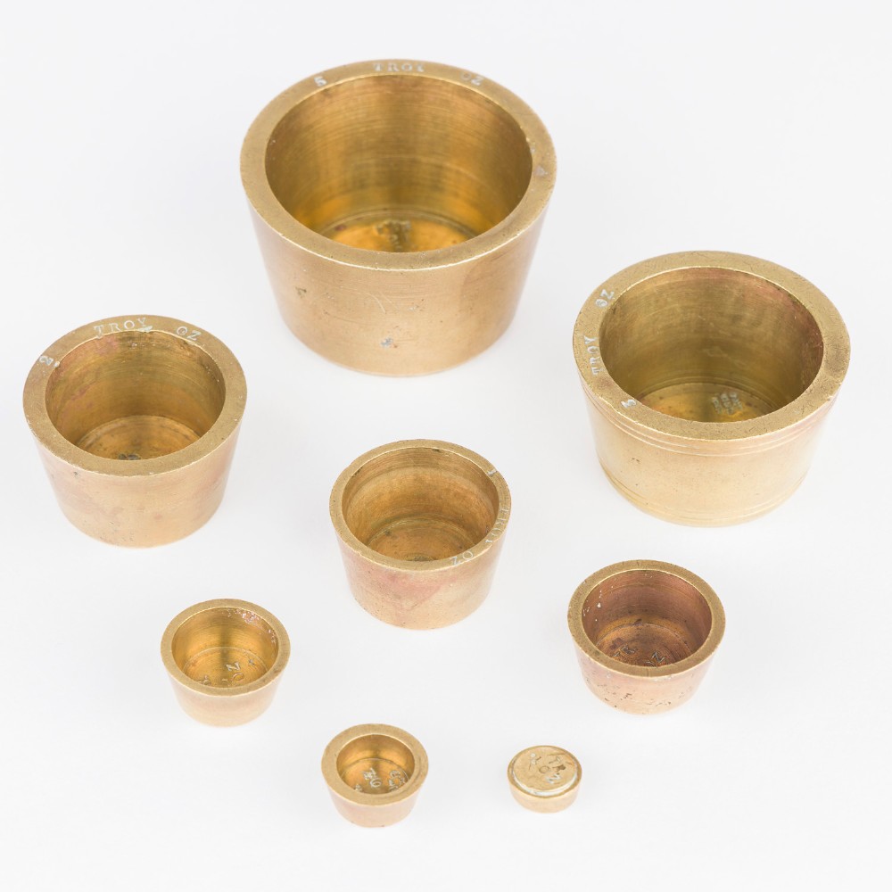 A set of eight graduated cylindrical brass nesting cup weights arranged in two loose rows on a plain white surface. View VI