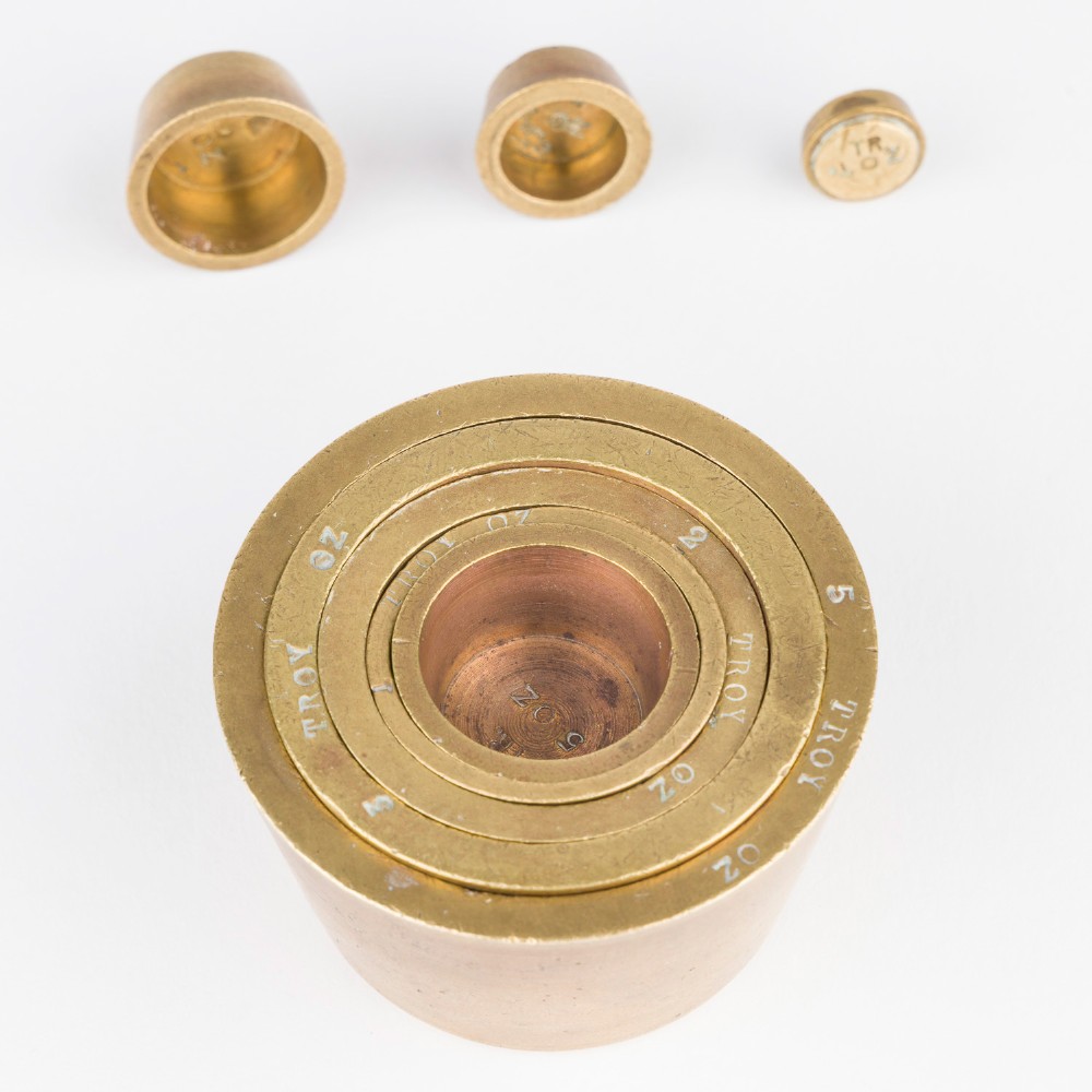 A set of eight graduated cylindrical brass nesting cup weights arranged in two loose rows on a plain white surface. View VII