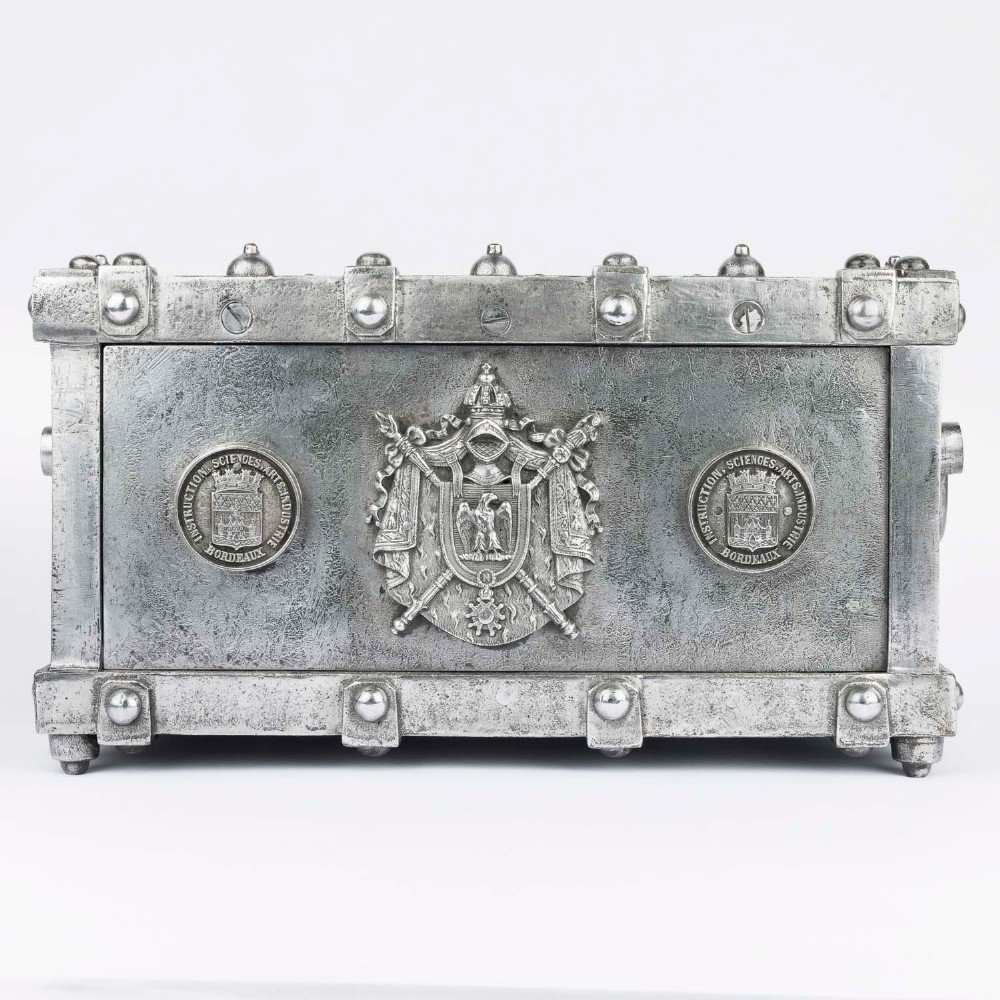 A heavy rectangular metal strongbox featuring riveted steel straps, raised studs, an embossed coat of arms, and maker plaques. View II
