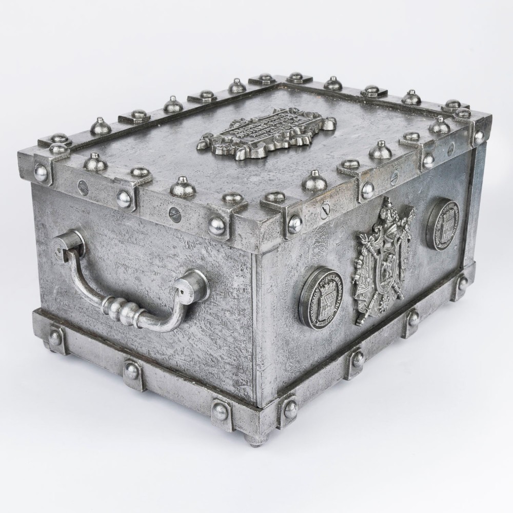A heavy rectangular metal strongbox featuring riveted steel straps, raised studs, an embossed coat of arms, and maker plaques. View III