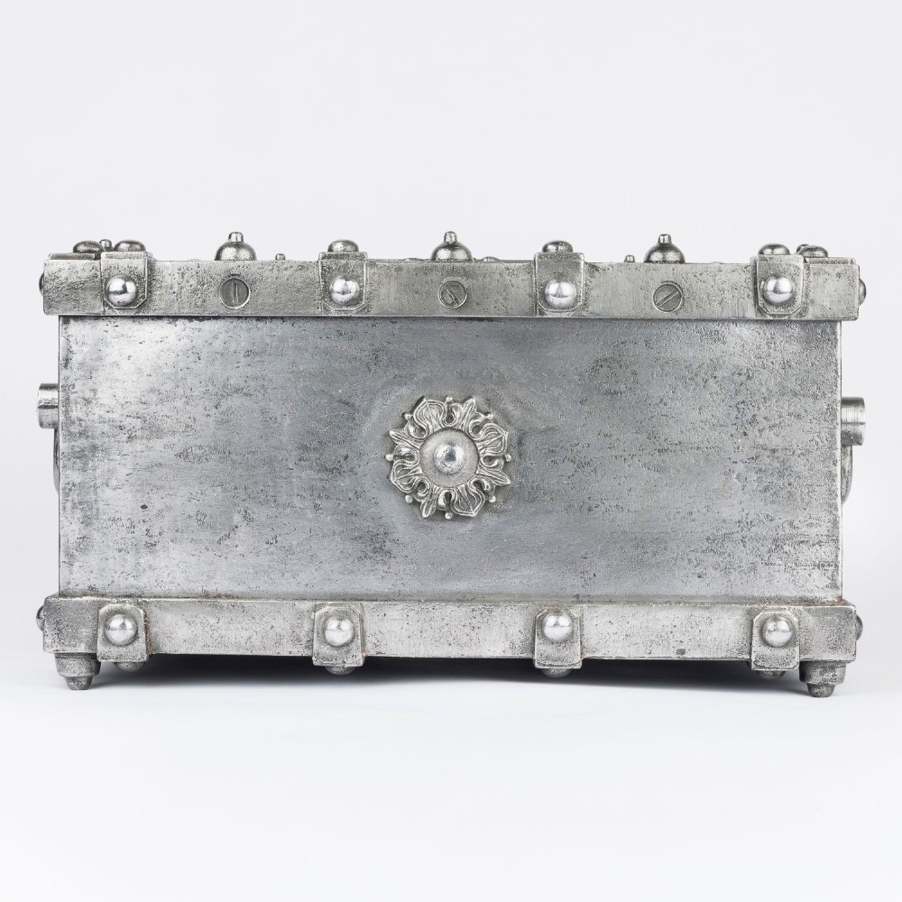 A heavy rectangular metal strongbox featuring riveted steel straps, raised studs, an embossed coat of arms, and maker plaques. View V
