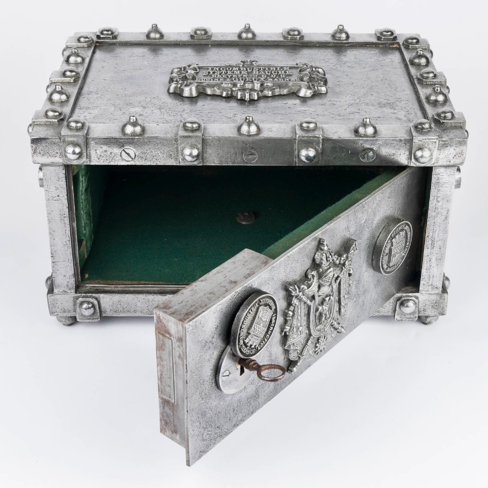 A heavy rectangular metal strongbox featuring riveted steel straps, raised studs, an embossed coat of arms, and maker plaques. View VI
