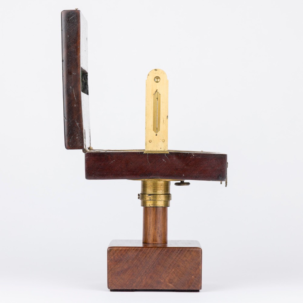 An antique mahogany cased surveyor compass with folding brass sight vanes and a circular glass-covered needle face, mounted on a wooden pedestal base. View V
