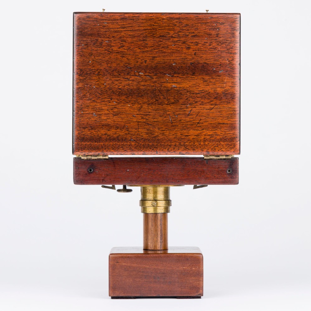 An antique mahogany cased surveyor compass with folding brass sight vanes and a circular glass-covered needle face, mounted on a wooden pedestal base. View X