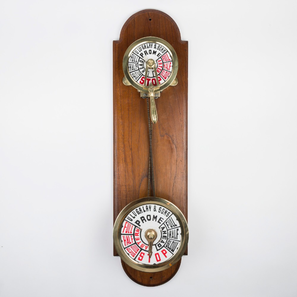 An antique brass engine room telegraph with two dials connected by a chain on a tall, arched timber backboard.
