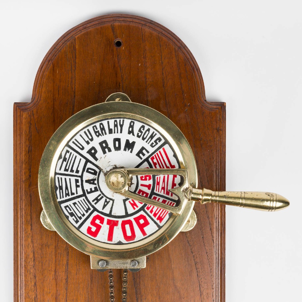 An antique brass engine room telegraph with two dials connected by a chain on a tall, arched timber backboard. View II