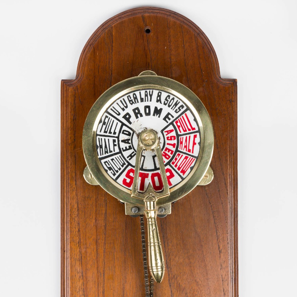An antique brass engine room telegraph with two dials connected by a chain on a tall, arched timber backboard. View III