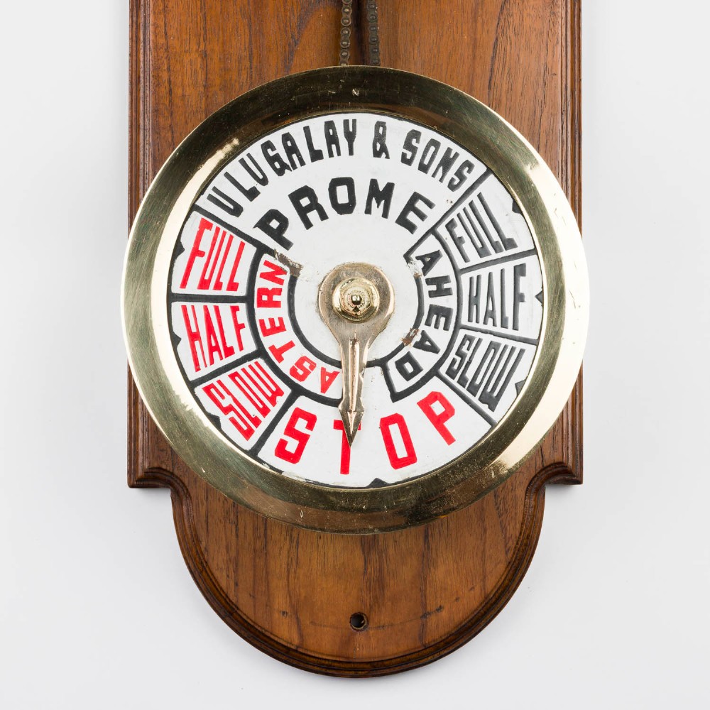 An antique brass engine room telegraph with two dials connected by a chain on a tall, arched timber backboard. View IV