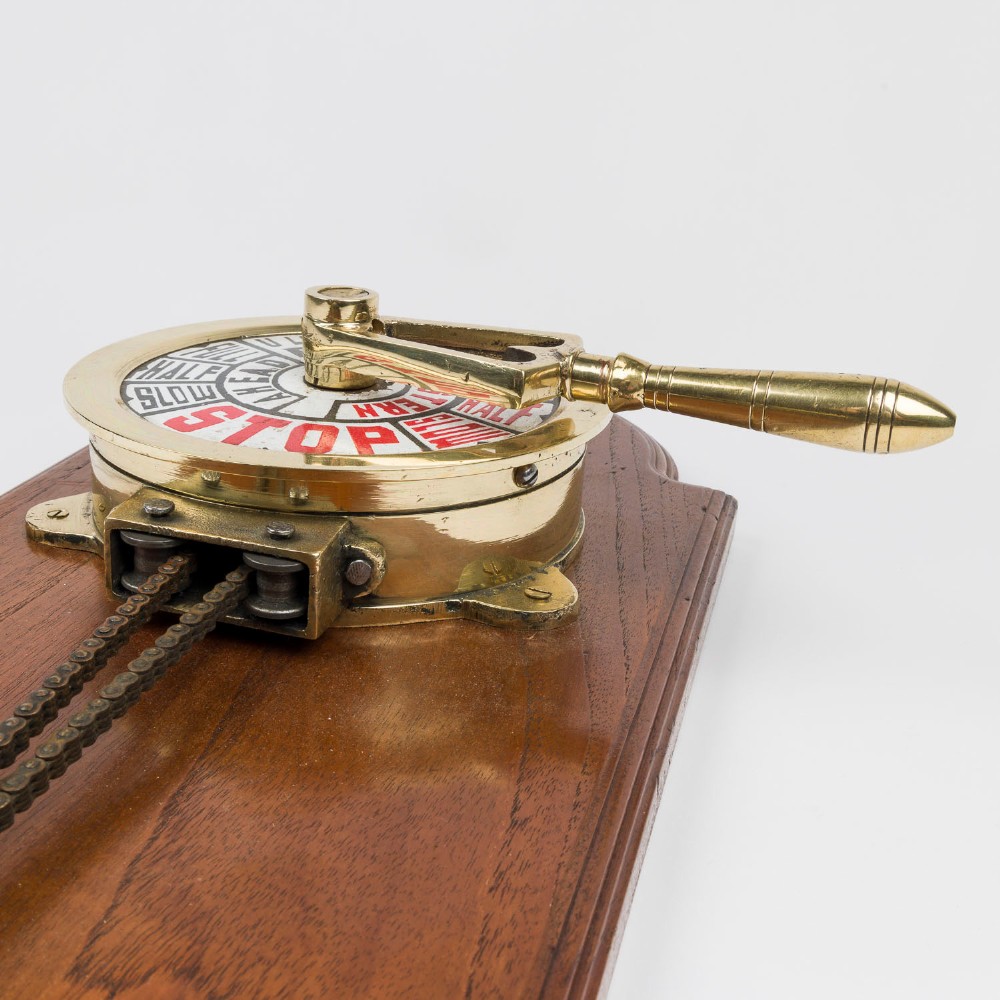 An antique brass engine room telegraph with two dials connected by a chain on a tall, arched timber backboard. View V