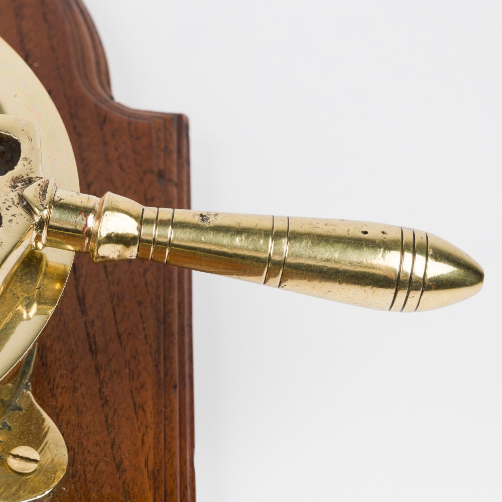 An antique brass engine room telegraph with two dials connected by a chain on a tall, arched timber backboard. View VI