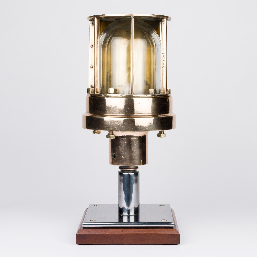 A polished brass submarine steaming light featuring a clear glass dome encased in a protective metal cage, mounted on a chrome stem and wooden plinth.