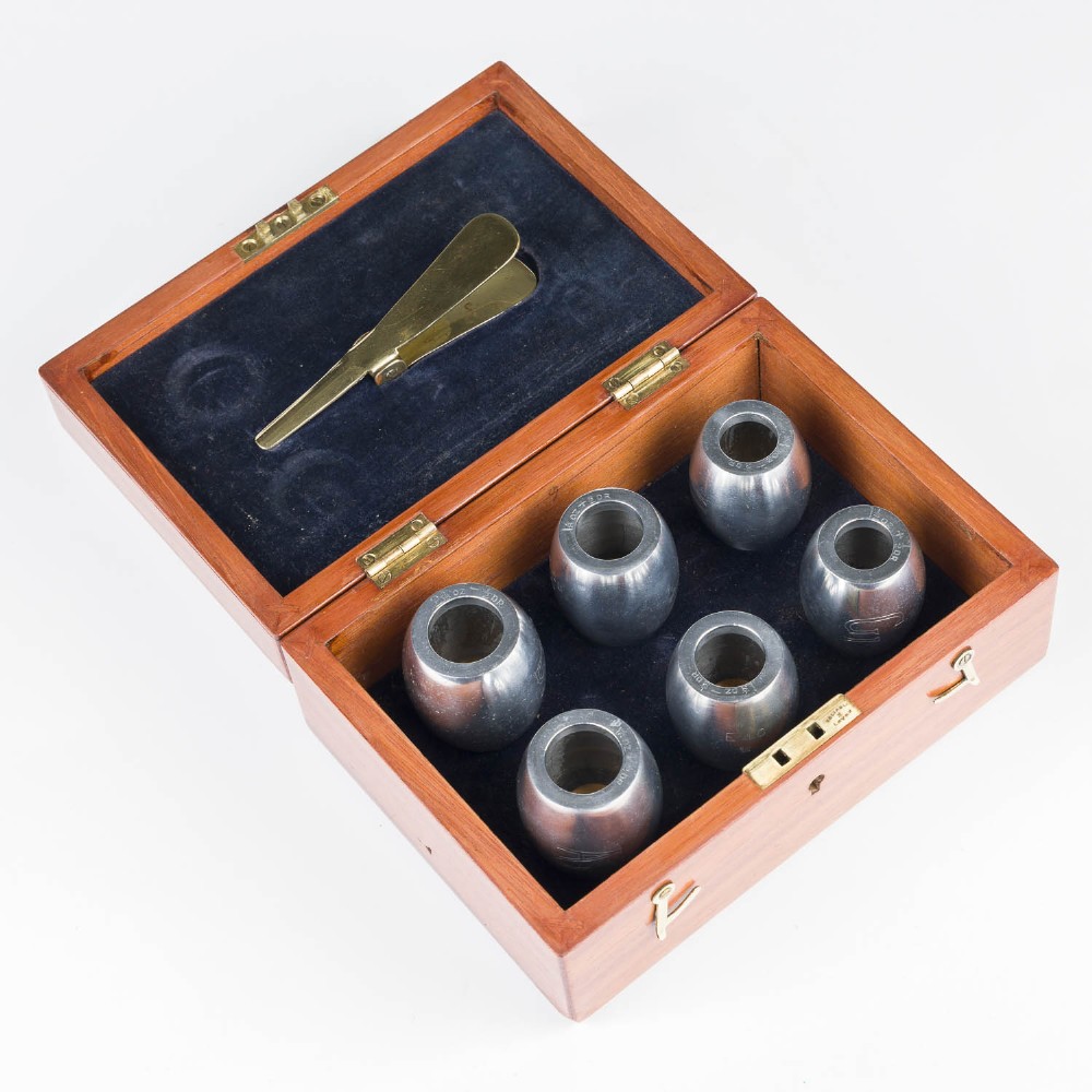 An open mahogany wooden box lined with blue velvet, containing six rounded metal egg weights and brass tweezers fitted into the lid.