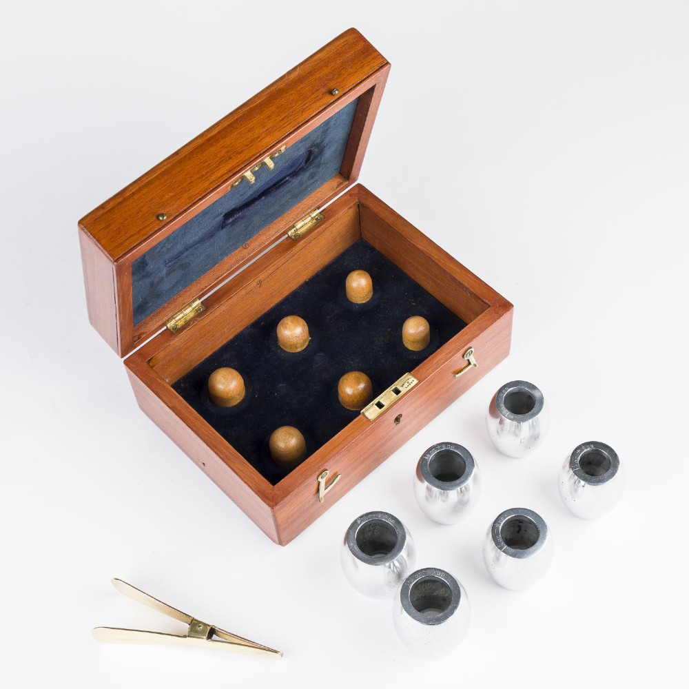 An open mahogany wooden box lined with blue velvet, containing six rounded metal egg weights and brass tweezers fitted into the lid. View II
