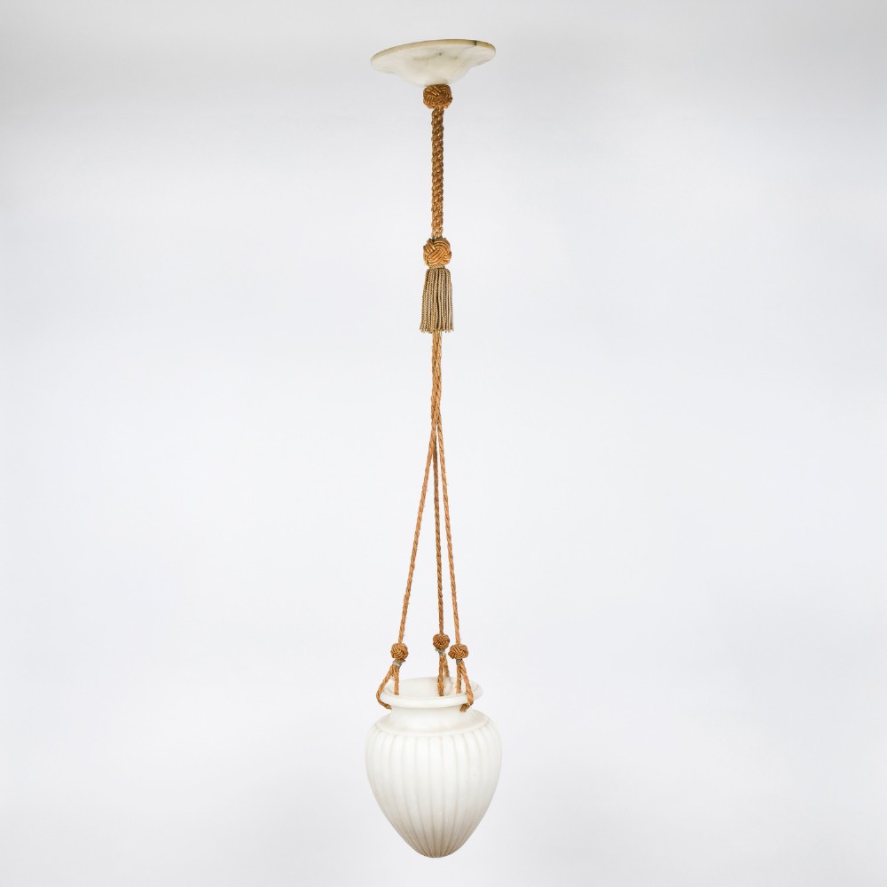 A fluted white alabaster urn-shaped light shade suspended by decorative woven cords and tassels from a matching alabaster ceiling rose.