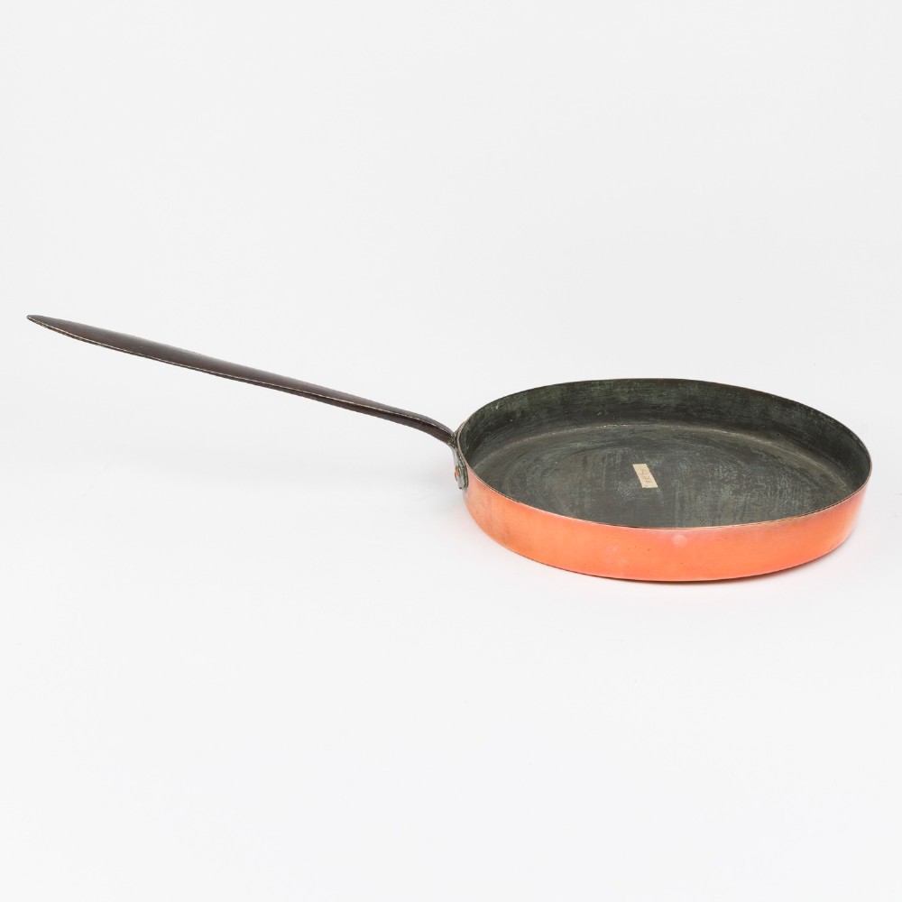 A circular antique copper saute pan with a dark tin-lined interior and a long dark iron handle.