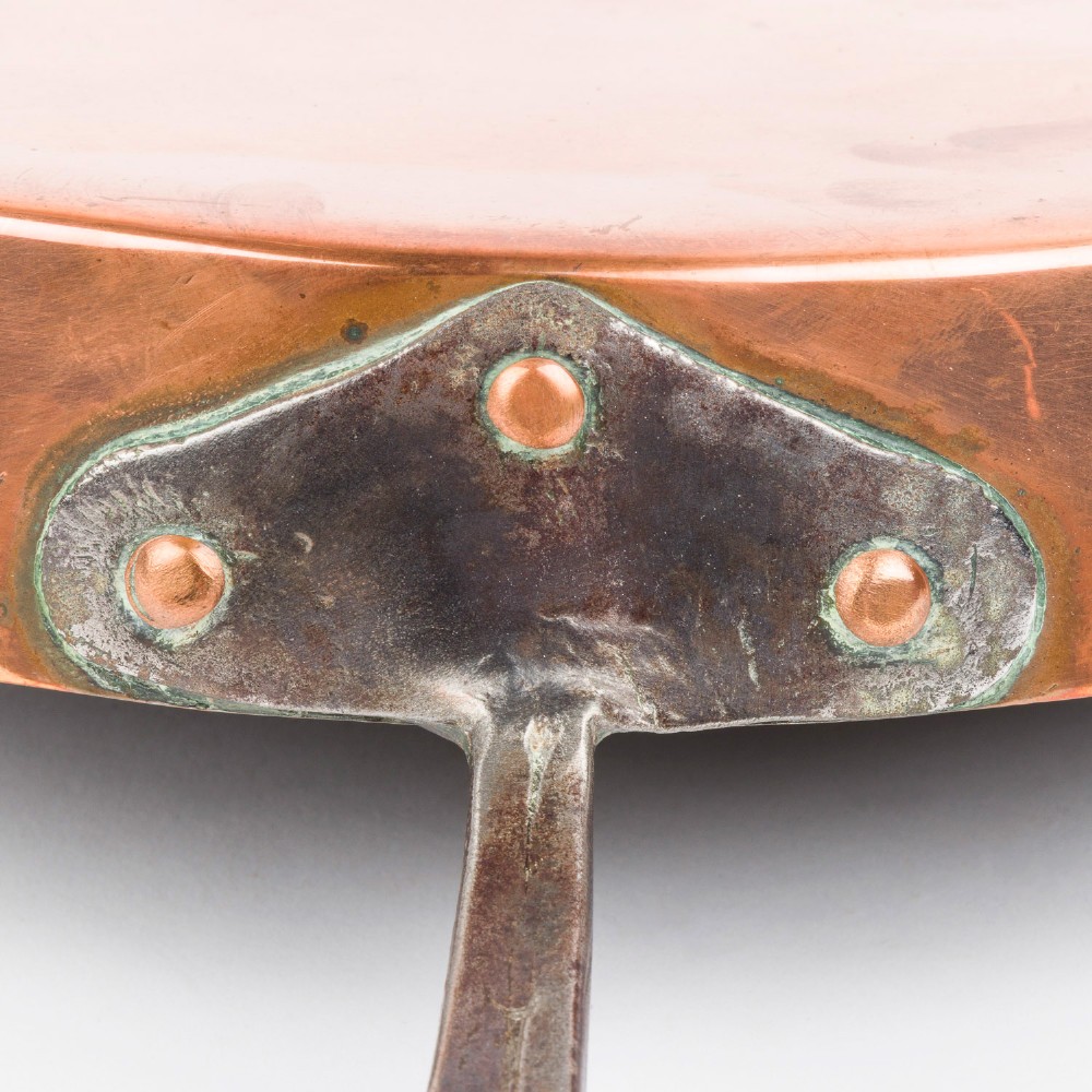 A circular antique copper saute pan with a dark tin-lined interior and a long dark iron handle. View IV