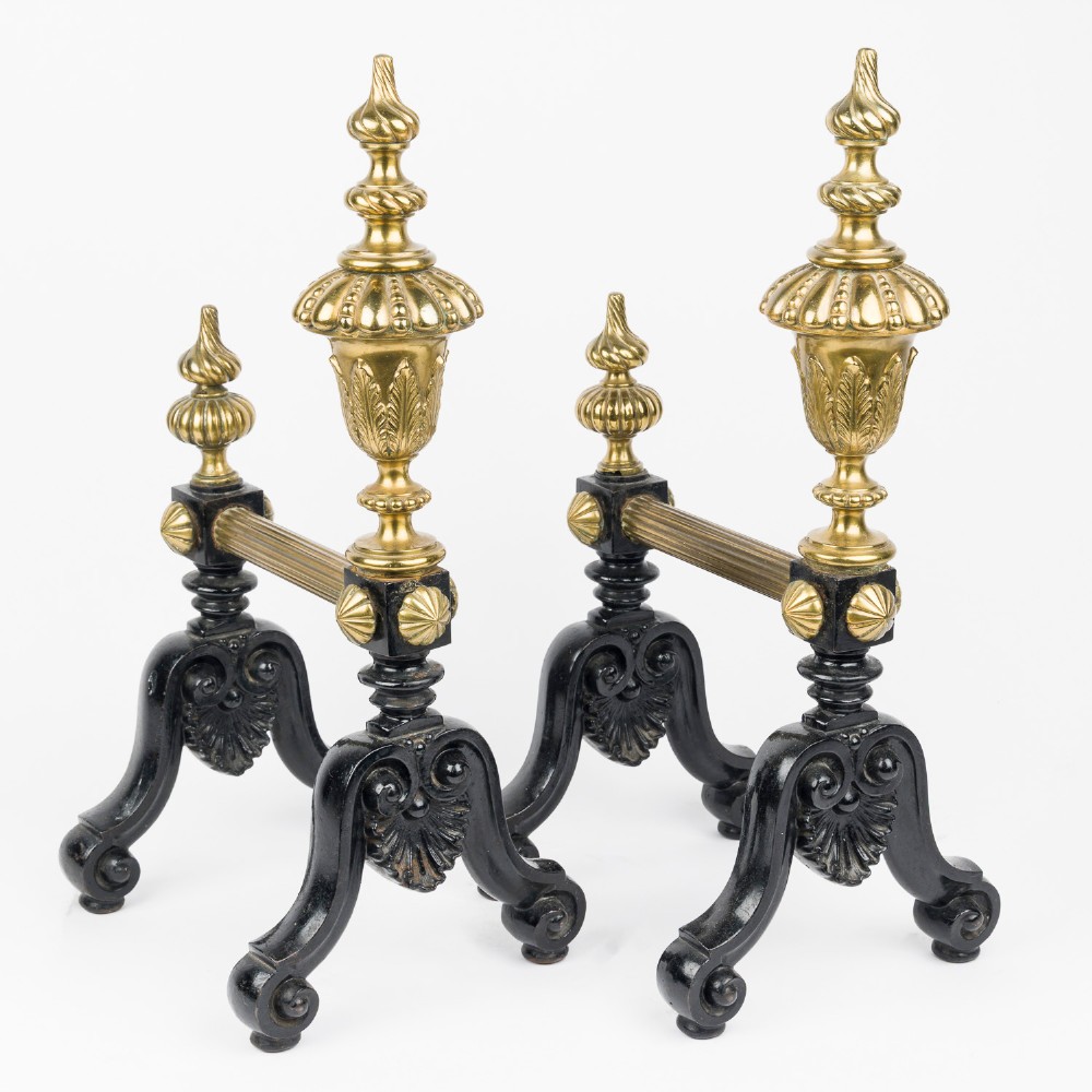 A pair of cast iron and brass fire dogs with ornate brass urn finials, fluted central bars, and black scroll feet.