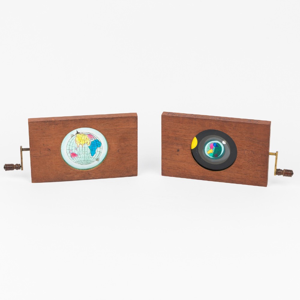 A pair of rectangular mahogany-framed mechanical magic lantern slides featuring hand-coloured astronomical glass apertures and brass hand cranks.
