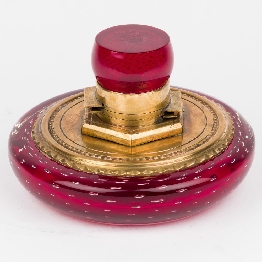 A circular ruby red glass inkwell featuring a controlled bubble pattern with a brass collar and matching glass stopper.