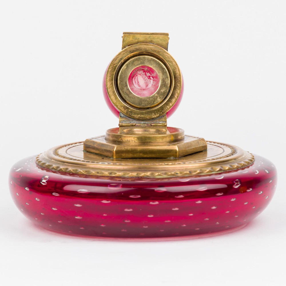 A circular ruby red glass inkwell featuring a controlled bubble pattern with a brass collar and matching glass stopper. View V