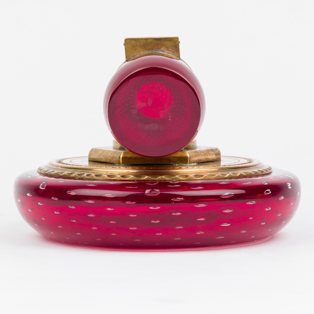 A circular ruby red glass inkwell featuring a controlled bubble pattern with a brass collar and matching glass stopper. View VI