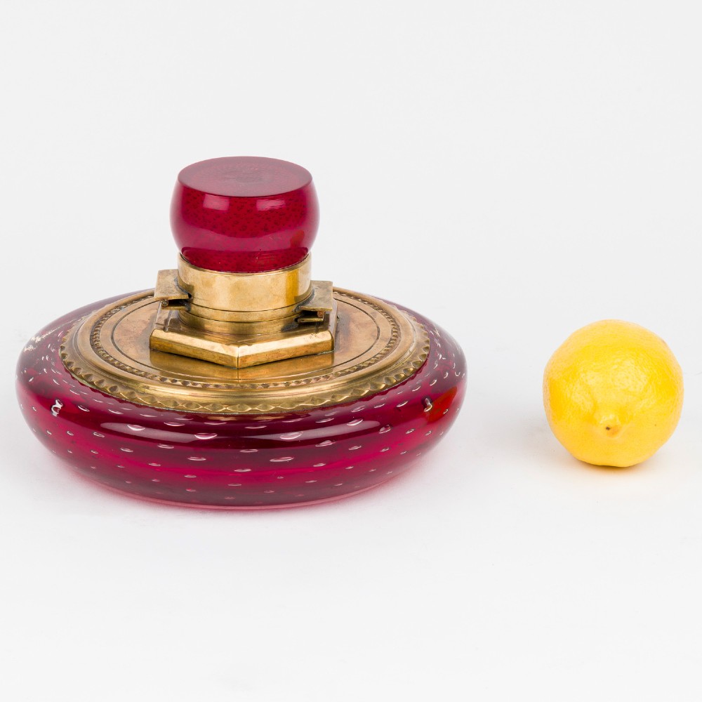 A circular ruby red glass inkwell featuring a controlled bubble pattern with a brass collar and matching glass stopper. View VIII