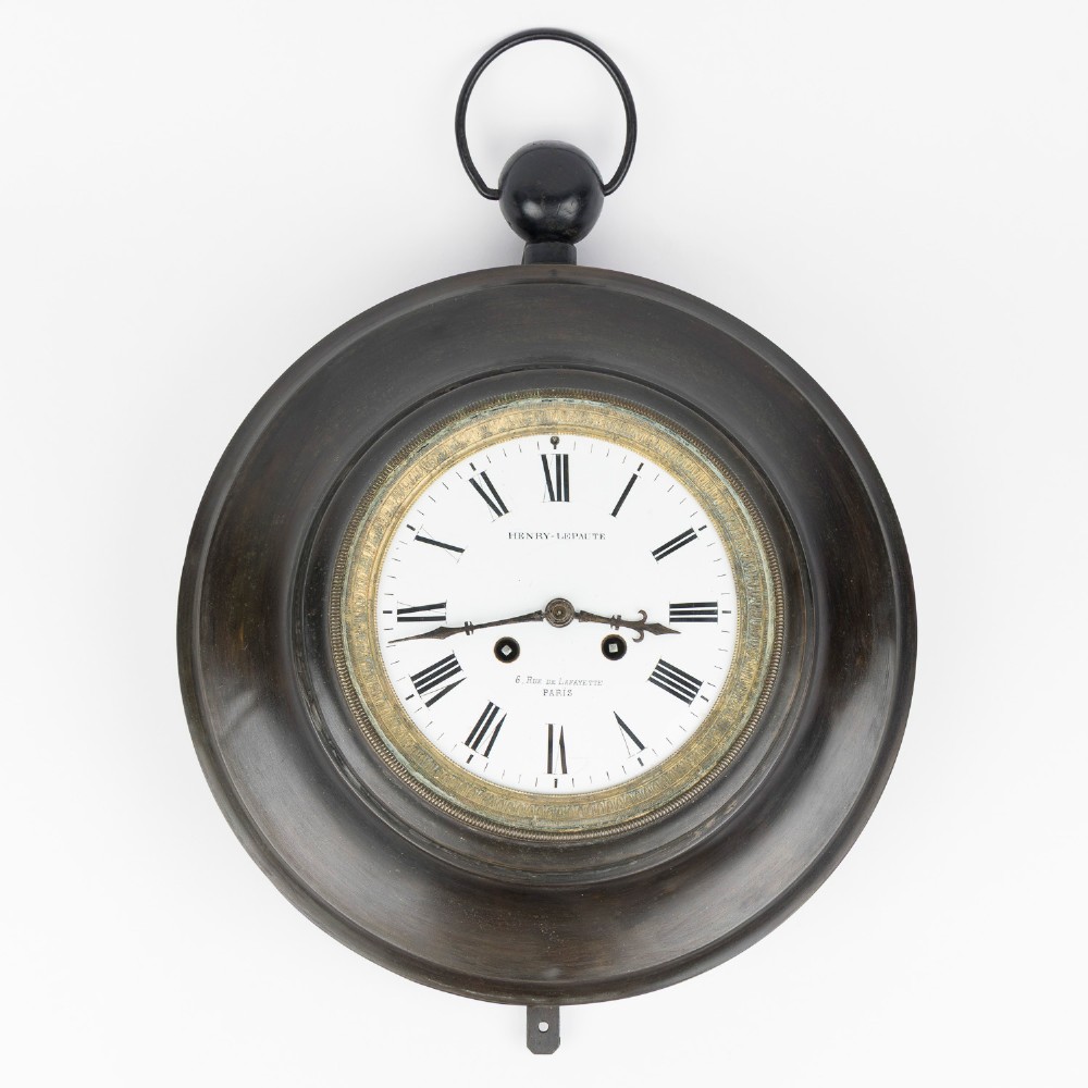 A circular dark bronze French wall clock designed in the shape of a pocket watch with a white enamel dial and Roman numerals.