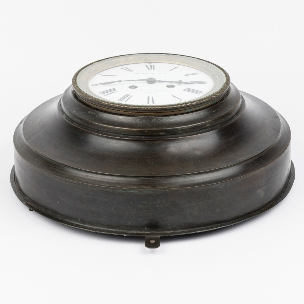A circular dark bronze French wall clock designed in the shape of a pocket watch with a white enamel dial and Roman numerals. View IV