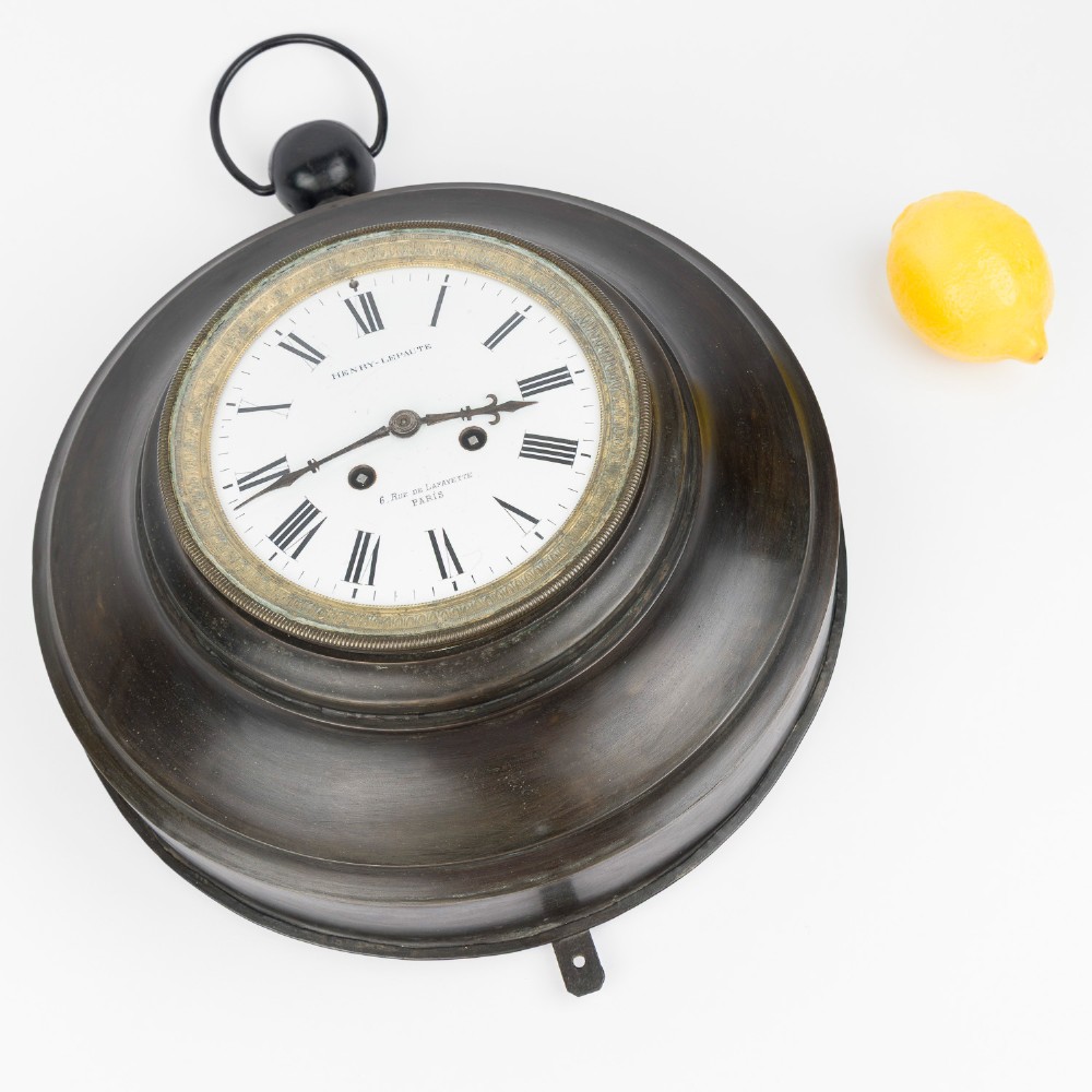 A circular dark bronze French wall clock designed in the shape of a pocket watch with a white enamel dial and Roman numerals. View XI