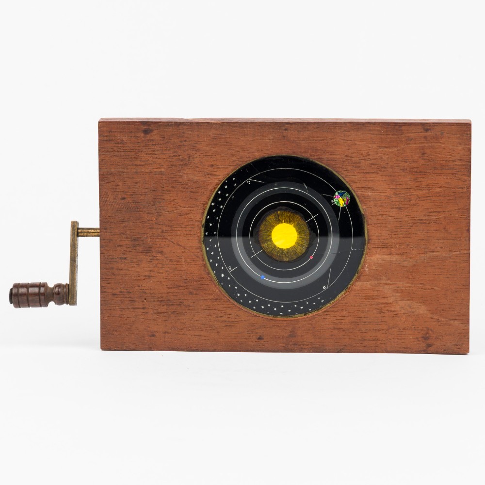 An antique mahogany wooden frame holding a circular hand-painted glass astronomical slide depicting planetary orbits around the sun, featuring a brass hand crank on the side.