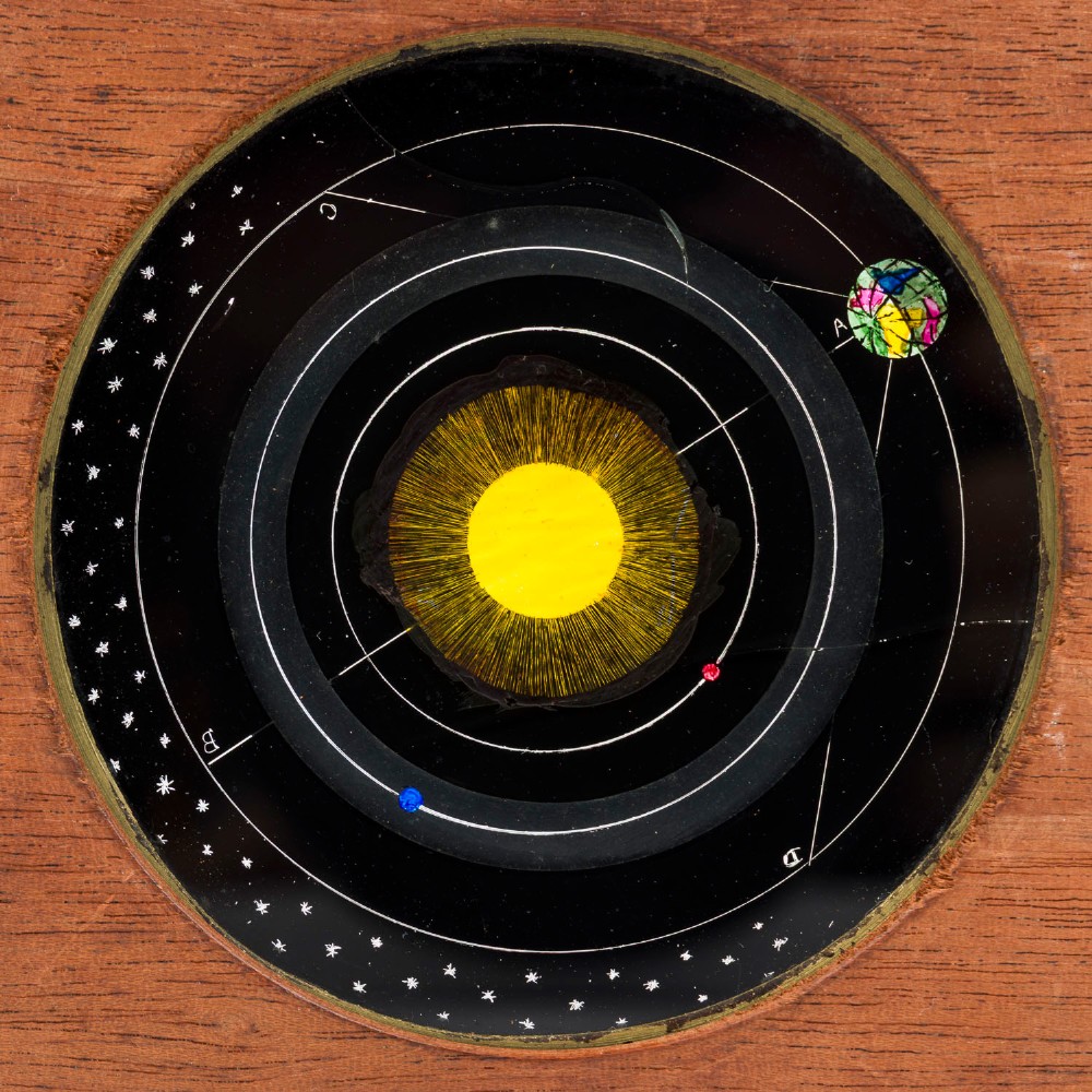 An antique mahogany wooden frame holding a circular hand-painted glass astronomical slide depicting planetary orbits around the sun, featuring a brass hand crank on the side. View II