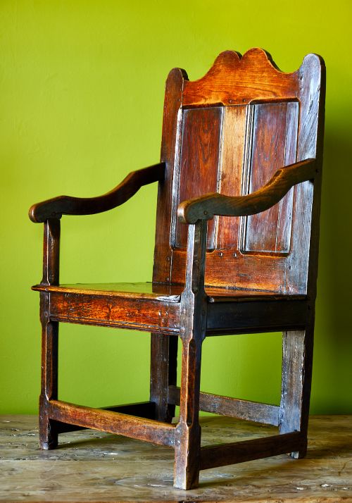 fig 2. A late 17th-century dark oak armchair featuring a panelled backrest, shaped crest rail, curved armrests, rectangular wooden seat, and square legs with low stretchers.