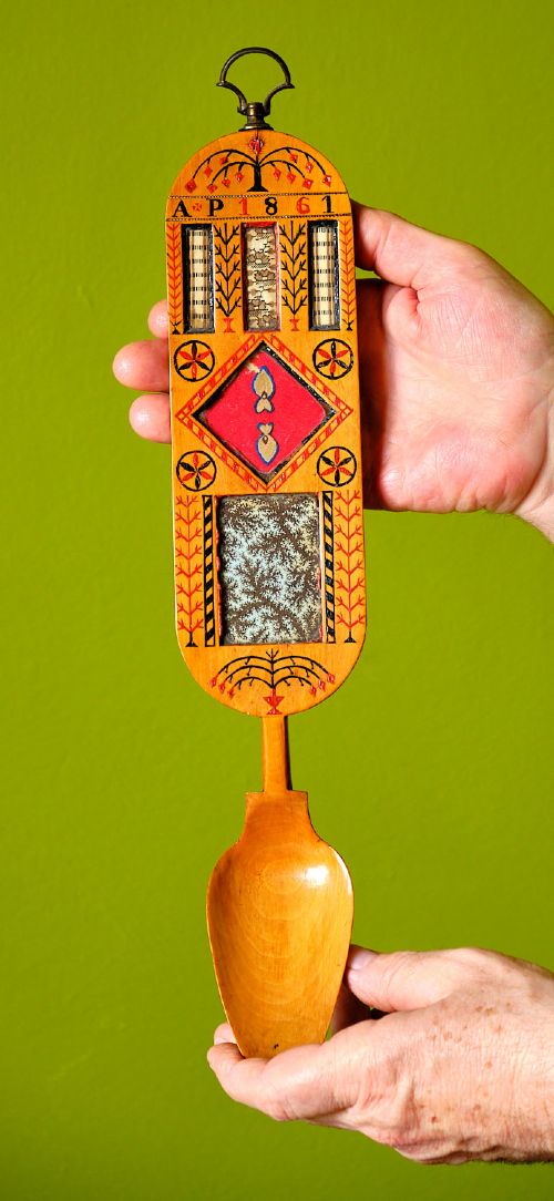A carved wooden Welsh love spoon featuring polychrome wax inlay, glazed inset panels, incised initials AP, the date 1861, and a brass suspension loop at the top. View VI