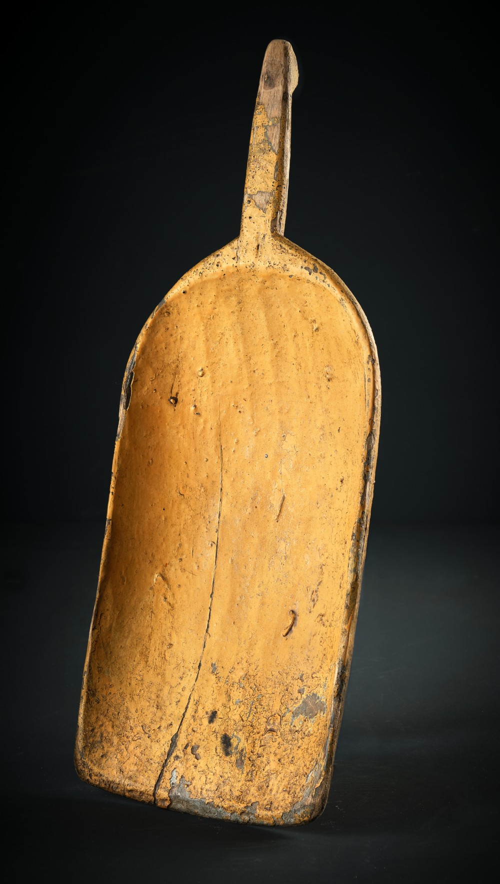 An antique carved wooden grain scoop featuring distressed yellow ochre paint and an integral shaped handle.