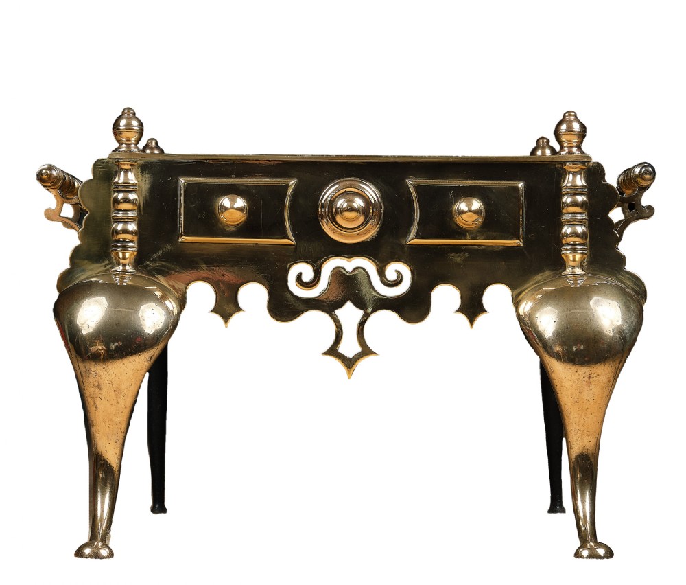 A polished brass Welsh footman hearth trivet with cabriole front legs, turned finials, carrying handles, and a pierced apron.