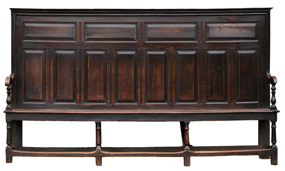 A high-backed dark oak settle with fielded panelling across the backrest, turned front legs, and a long wooden seat.