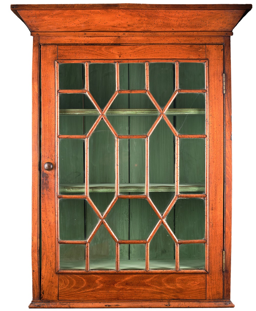 An antique timber wall cabinet featuring a geometric glazed glass door and a painted green interior with shelves.