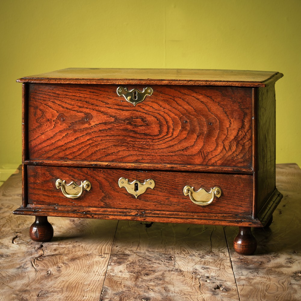An 18th-century Welsh oak coffor bach featuring a rich wood grain, a hinged top section, a lower drawer with brass handles, and turned feet.