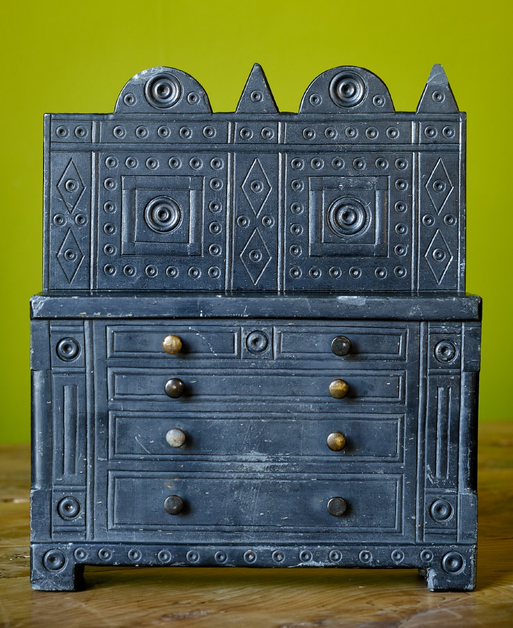 A miniature carved dark slate chest of drawers featuring geometric engravings, circular motifs, and small brass knobs.