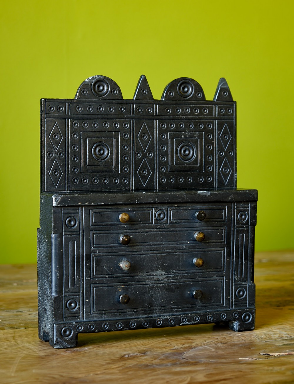 A miniature carved dark slate chest of drawers featuring geometric engravings, circular motifs, and small brass knobs. View II