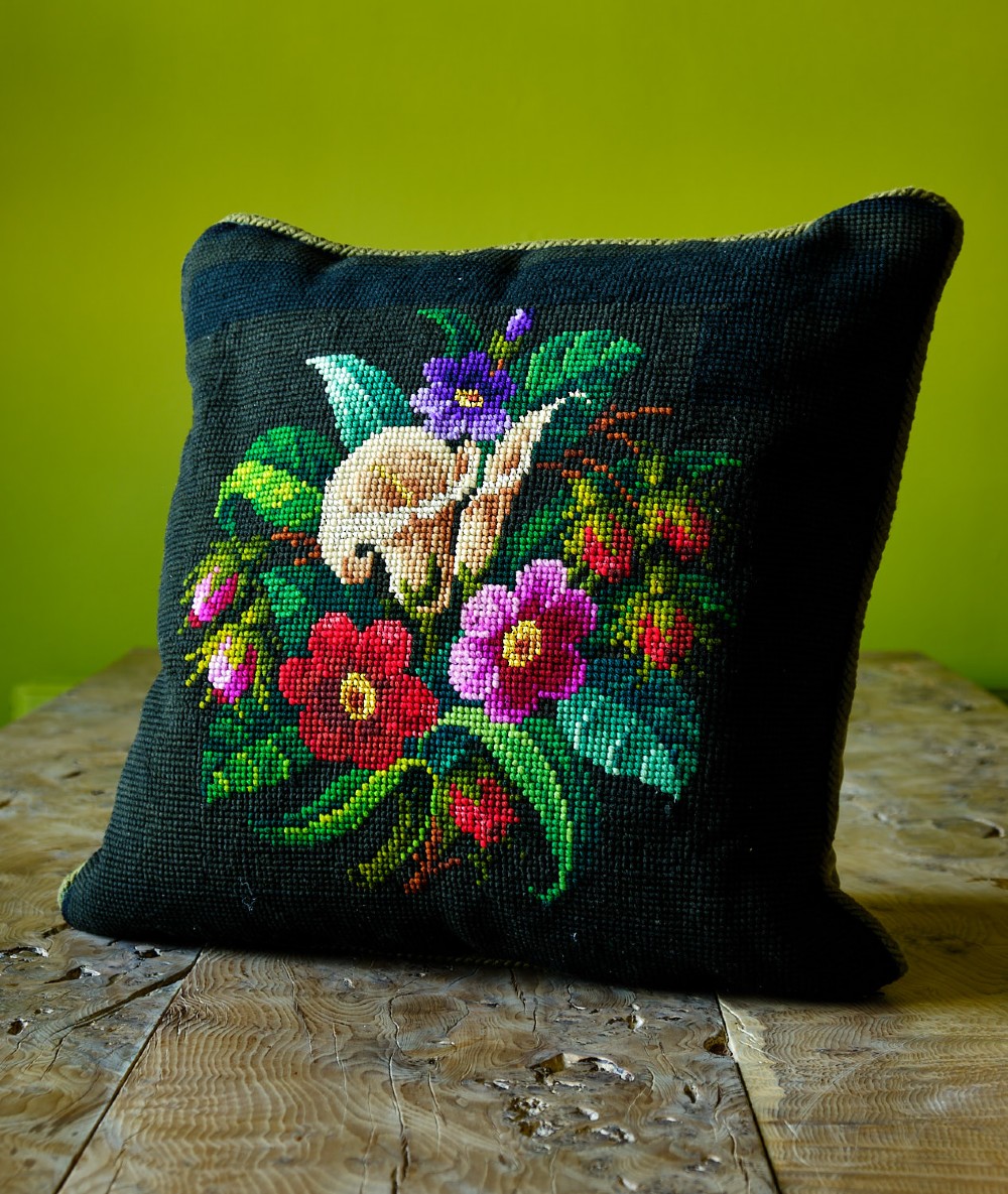 A square dark cushion featuring a central hand-stitched Victorian woolwork and silkwork floral motif with red, pink, and purple flowers.