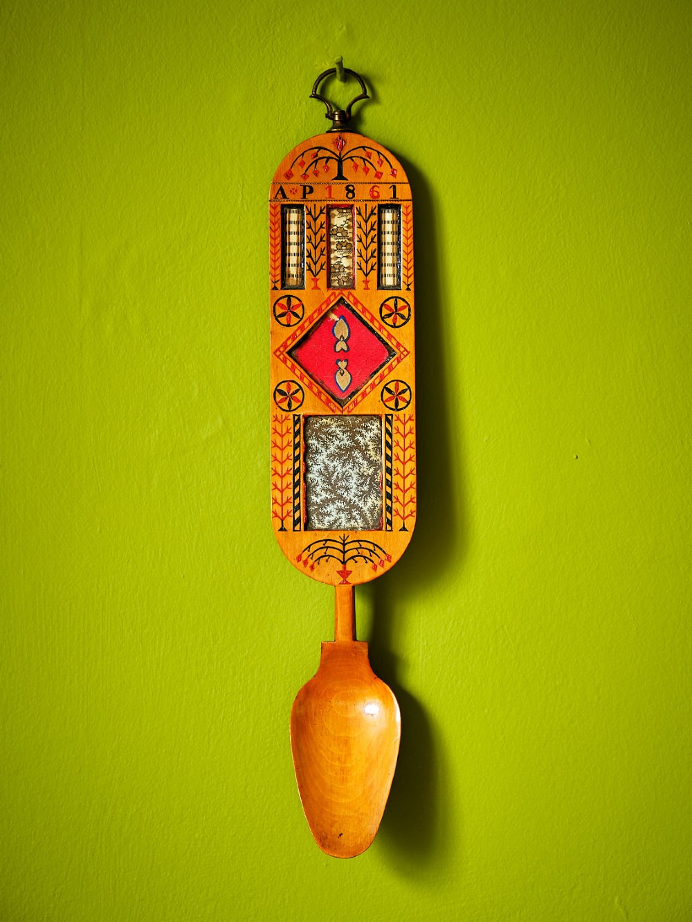 A carved wooden Welsh love spoon featuring polychrome wax inlay, glazed inset panels, incised initials AP, the date 1861, and a brass suspension loop at the top.