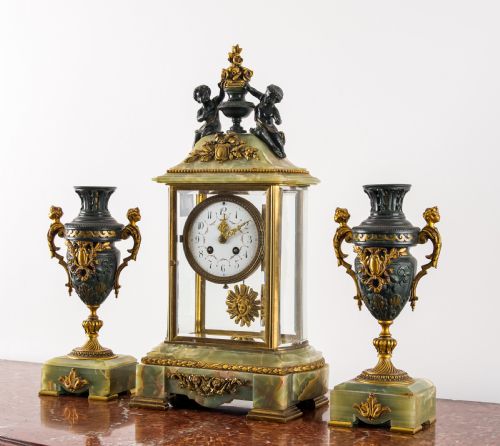 fig 1. A French three-piece garniture set featuring a central glazed onyx clock topped with dark bronze cherub figures, flanked by two matching side urns on green onyx bases.