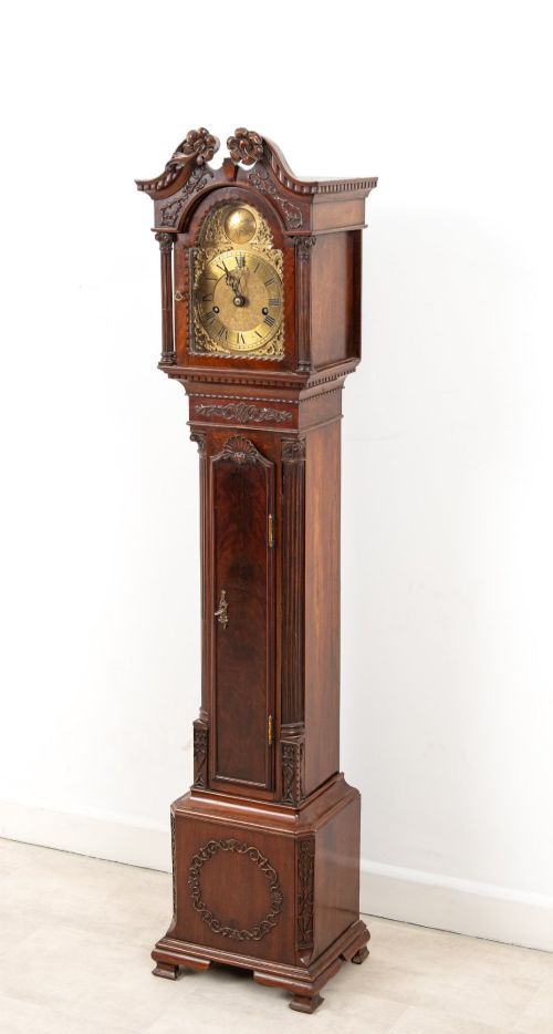 fig 2. An Edwardian mahogany grandmother clock with a carved swan neck pediment, a brass dial with Roman numerals, and a slim waist door.