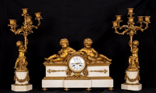 fig 1. A 19th-century French clock garniture featuring a central white marble and gilt bronze clock topped with two reclining cherub figures, flanked by a pair of matching five-light gilt bronze candelabra supported by cherubs on marble bases.
