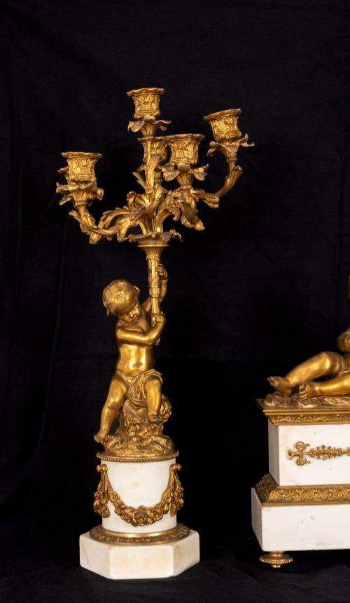 fig 2. A 19th-century French clock garniture featuring a central white marble and gilt bronze clock topped with two reclining cherub figures, flanked by a pair of matching five-light gilt bronze candelabra supported by cherubs on marble bases.