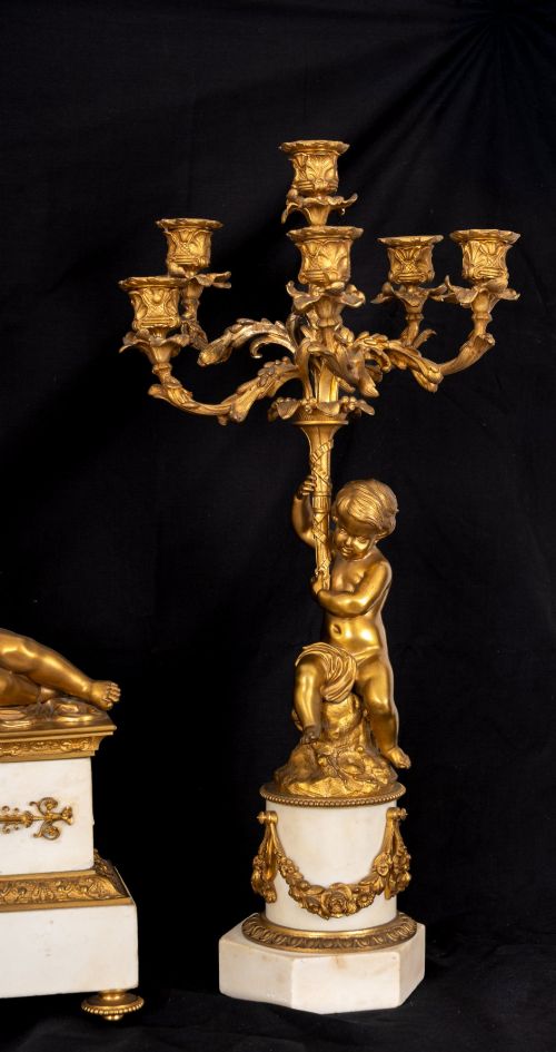fig 3. A 19th-century French clock garniture featuring a central white marble and gilt bronze clock topped with two reclining cherub figures, flanked by a pair of matching five-light gilt bronze candelabra supported by cherubs on marble bases.